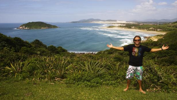 Ein Mann steht mit ausgebreiteten Armen vor einer Küstenlandschaft mit Insel und Strand.