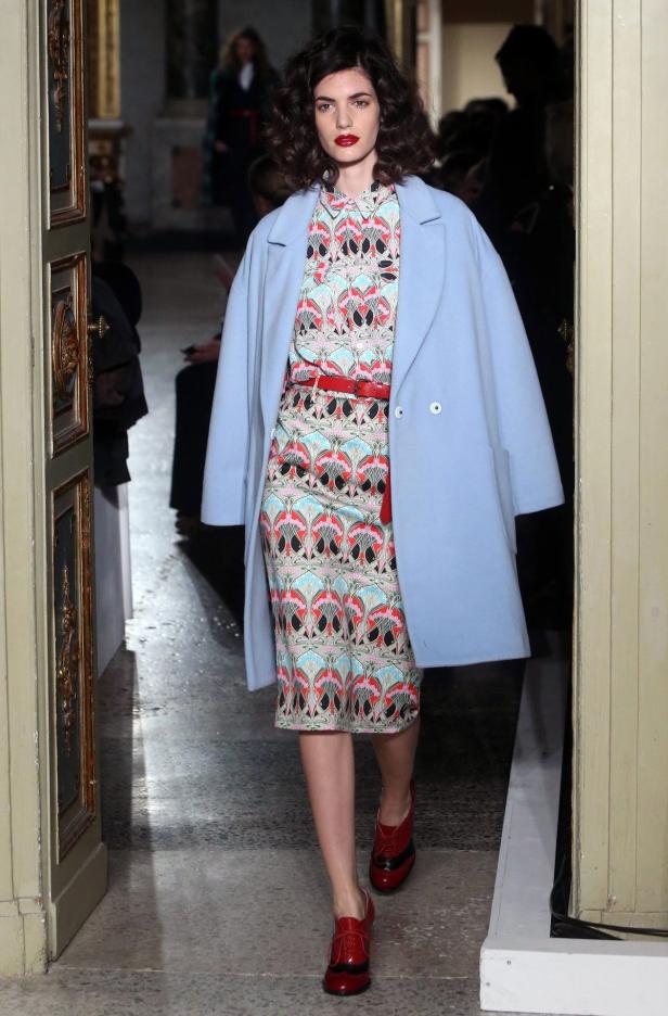 Eine Frau mit einem gemusterten Kleid, einem hellblauen Mantel und roten Schuhen betritt einen Raum.