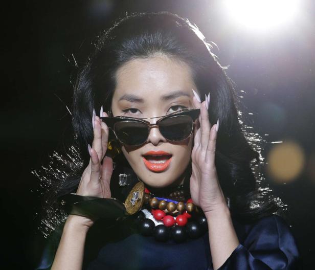 Eine Frau mit Sonnenbrille, auffälligem Schmuck und orangefarbenem Lippenstift blickt in die Kamera.