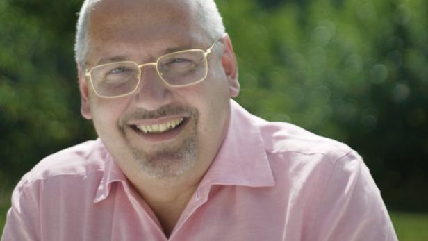 Ein lächelnder Mann mit Brille und rosa Hemd vor grünem Hintergrund.