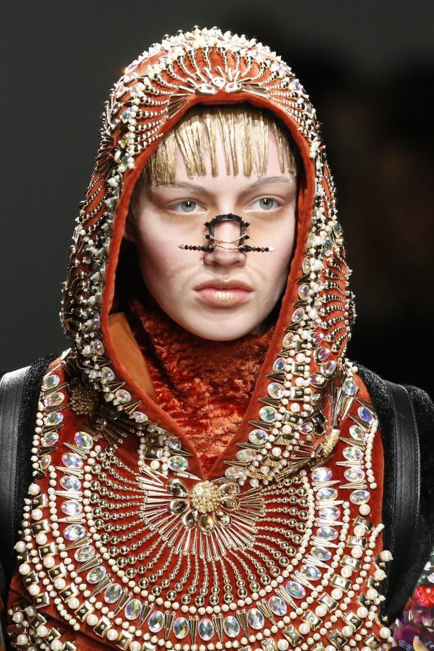 Ein Model mit goldenem Haar und Gesichtsschmuck trägt eine reich verzierte Kapuzenrobe.