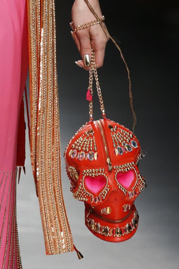 Eine rote, schädelgeformte Handtasche, verziert mit Nieten und Strasssteinen, wird von einer Hand gehalten.