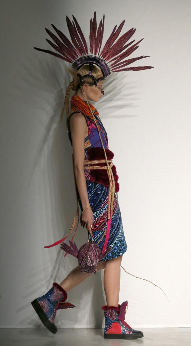Eine Frau in einem extravaganten Federkopfschmuck und farbenfrohem Kleid läuft an einer Wand entlang.