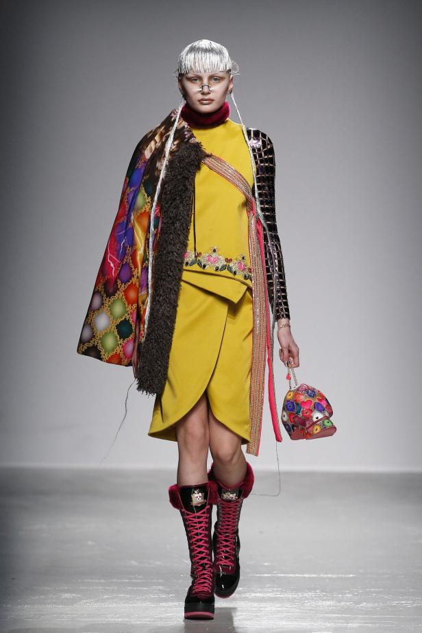 Eine Frau präsentiert ein gelbes Kleid, bunte Accessoires und hohe, pinkfarbene Stiefel auf einem Laufsteg.