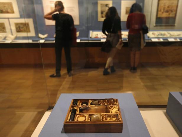Eine Holzkiste mit verschiedenen Objekten in einem Museum, im Hintergrund Besucher.