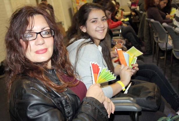 Zwei Frauen sitzen in einem Publikum und halten bunte Papierfächer in den Händen.