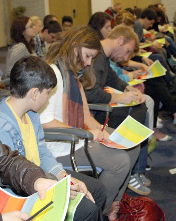 Eine Gruppe von Menschen sitzt in Stuhlreihen und füllt Fragebögen aus.
