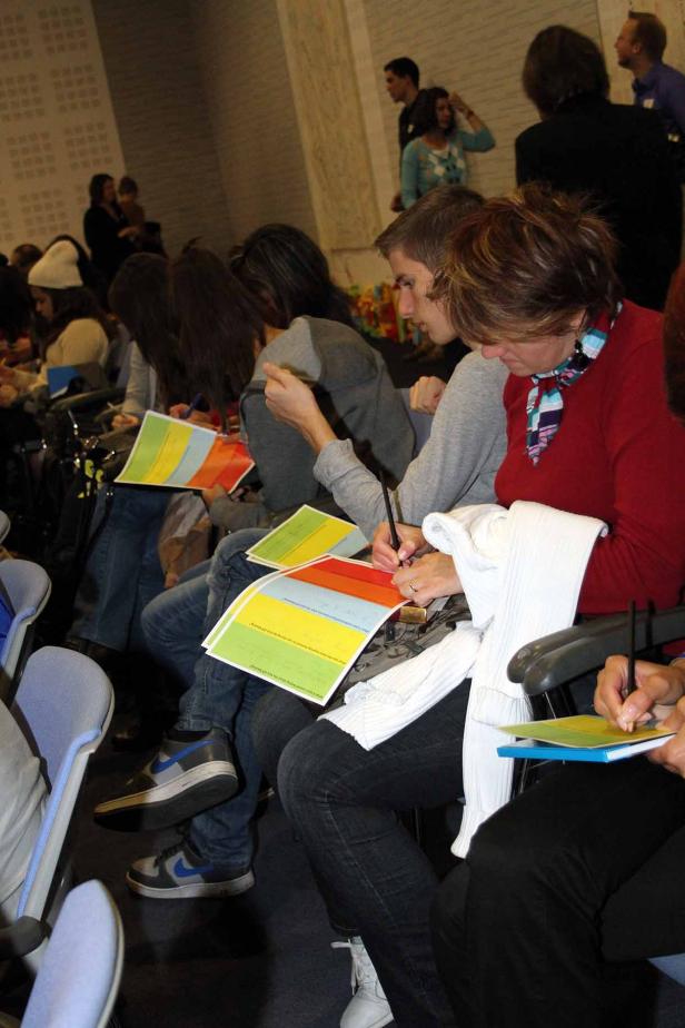 Menschen sitzen in einer Reihe und füllen farbige Fragebögen aus.