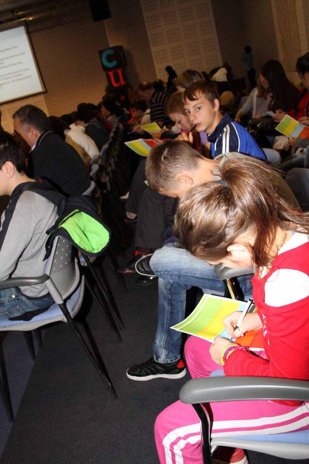 Kinder sitzen in einem Publikum und füllen farbige Blätter aus.