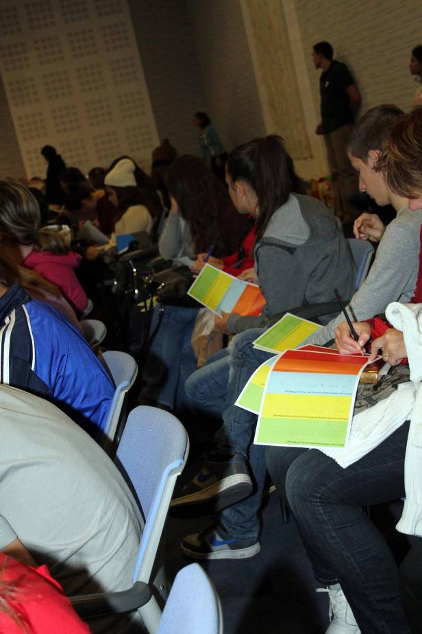 Mehrere Personen sitzen in einem Raum und füllen farbige Fragebögen aus.