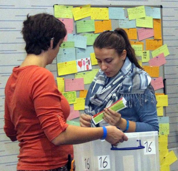 Zwei Frauen sortieren Zettel vor einer Wand voller Notizen.