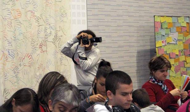 Eine Person fotografiert mit einer Kamera vor einer Wand mit Handabdrücken, während andere Personen im Raum sitzen.