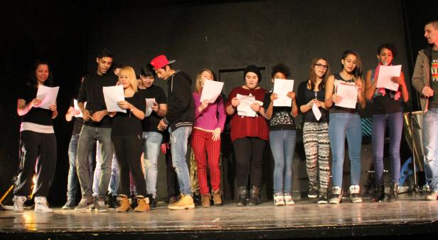 Eine Gruppe junger Leute steht auf einer Bühne und liest von Papierblättern.