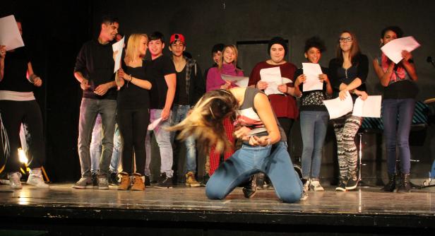 Eine junge Frau kniet auf der Bühne, während eine Gruppe von Menschen im Hintergrund steht und Blätter Papier hält.