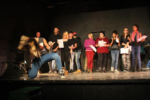 Eine Gruppe junger Leute steht auf einer Bühne, während eine Frau im Vordergrund tanzt.