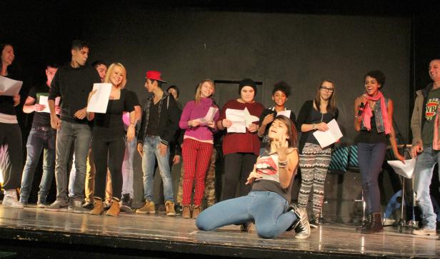 Eine Gruppe junger Leute steht auf einer Bühne, während ein Mädchen im Vordergrund kniet und gestikuliert.