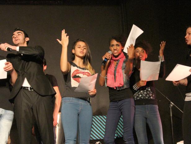 Eine Gruppe junger Leute steht auf einer Bühne und liest von Zetteln ab.