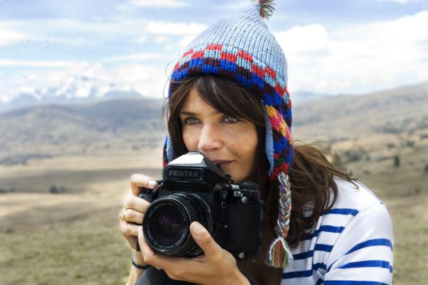 Eine Frau mit Mütze fotografiert mit einer Pentax-Kamera in einer hügeligen Landschaft.