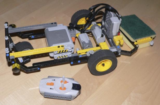 Ein Lego-Roboter mit Fernbedienung und einem Schwamm befestigt an der Rückseite steht auf einem Holzboden.