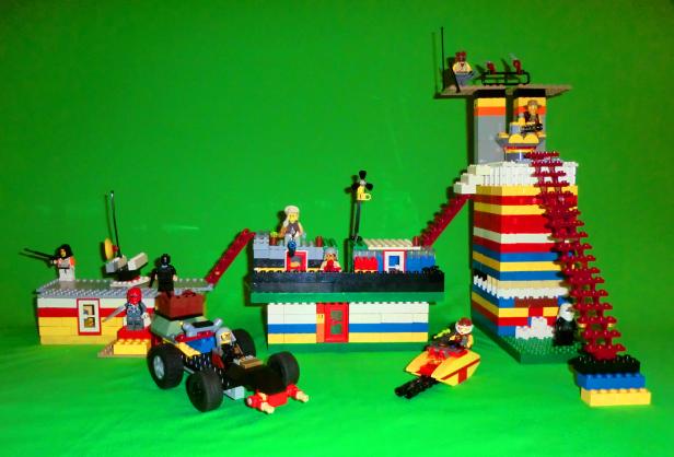 Eine Lego-Szene mit Figuren, Gebäuden und Fahrzeugen vor einem grünen Hintergrund.