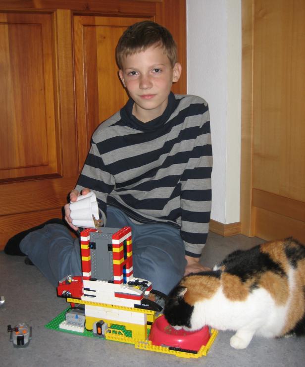 Ein Junge gießt Futter in ein Lego-Gebäude, während eine Katze aus einem roten Napf frisst.