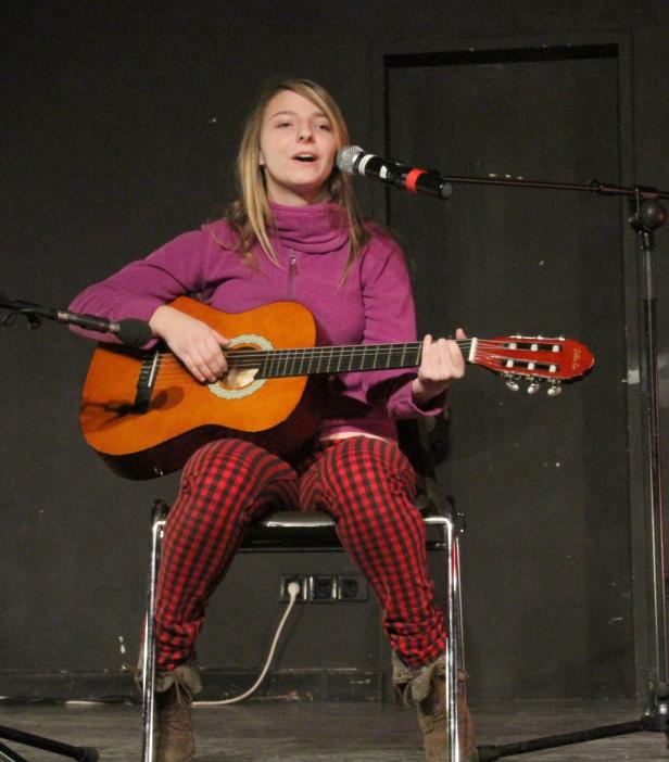 Eine junge Frau sitzt auf einem Stuhl, spielt Gitarre und singt in ein Mikrofon.