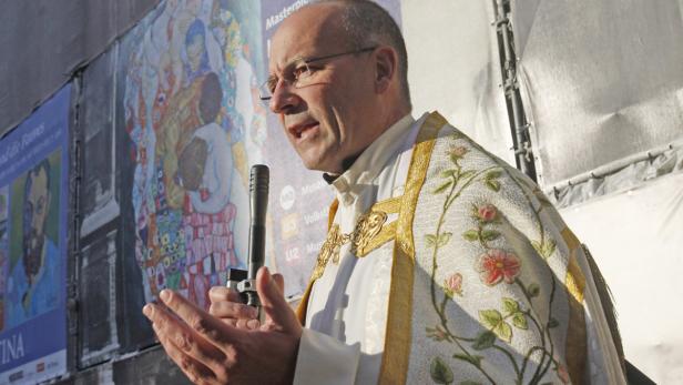 Ein Priester spricht vor einer Wand mit Kunstplakaten.
