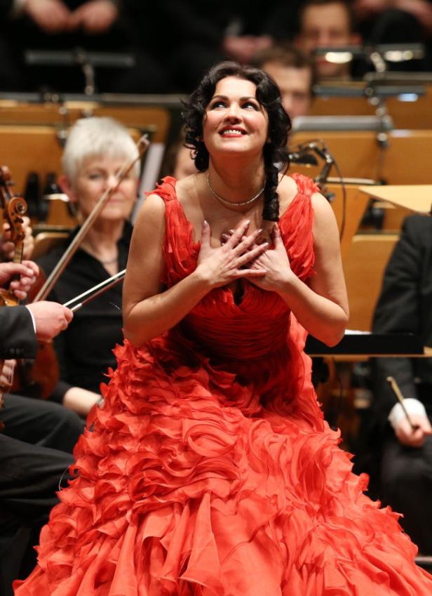 Eine Opernsängerin in einem roten Kleid mit Rüschen singt vor einem Orchester.
