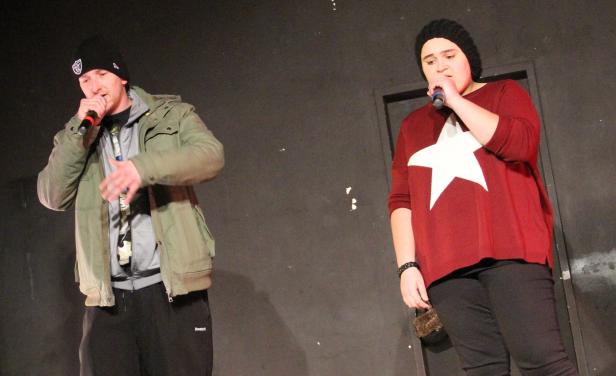 Zwei Personen mit Mützen stehen auf einer Bühne und singen in Mikrofone.