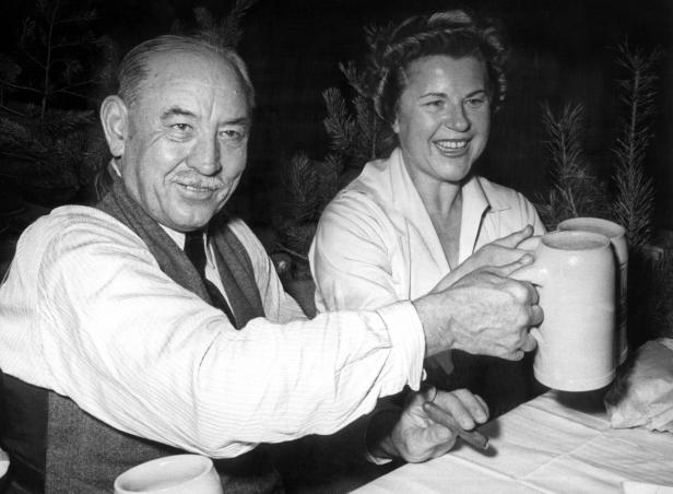 Ein Mann und eine Frau stoßen mit Bierkrügen an, der Mann raucht eine Zigarre.