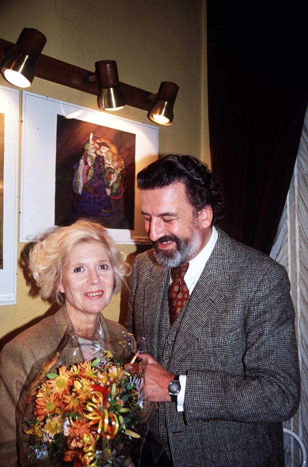 Eine Frau mit einem Blumenstrauß und ein Mann in einem karierten Anzug stehen zusammen vor einem Bild.