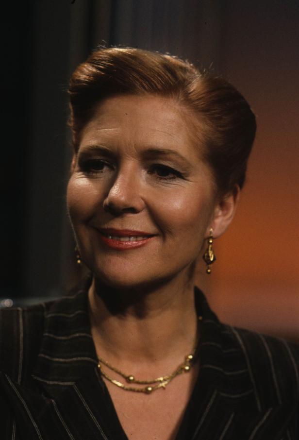 Eine lächelnde Frau mit rotem Haar, Ohrringen und einer goldenen Halskette.