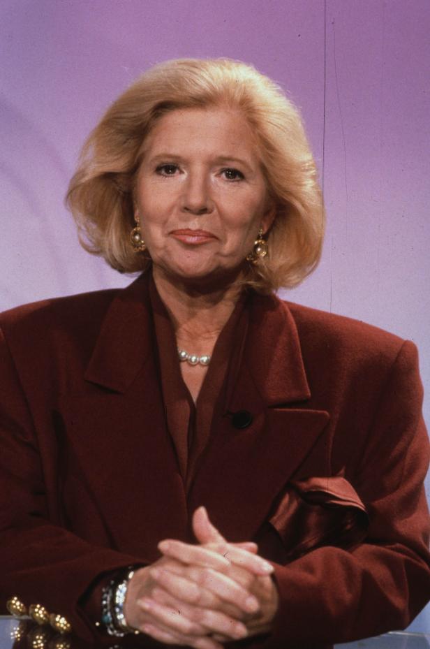 Eine Frau mit blonden Haaren, Perlenkette und rotem Blazer vor einem lila Hintergrund.