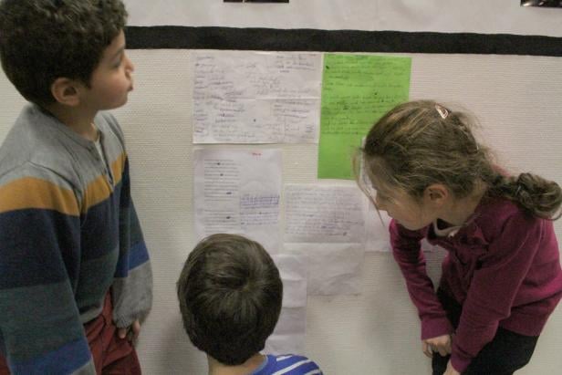 Drei Kinder betrachten beschriebene Blätter an einer Wand.