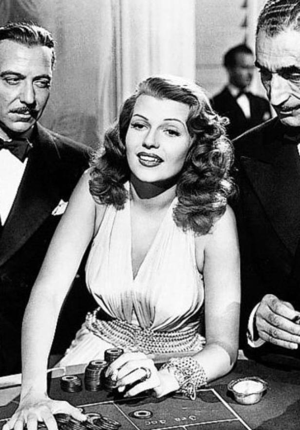 Rita Hayworth beim Glücksspiel, flankiert von zwei Männern.