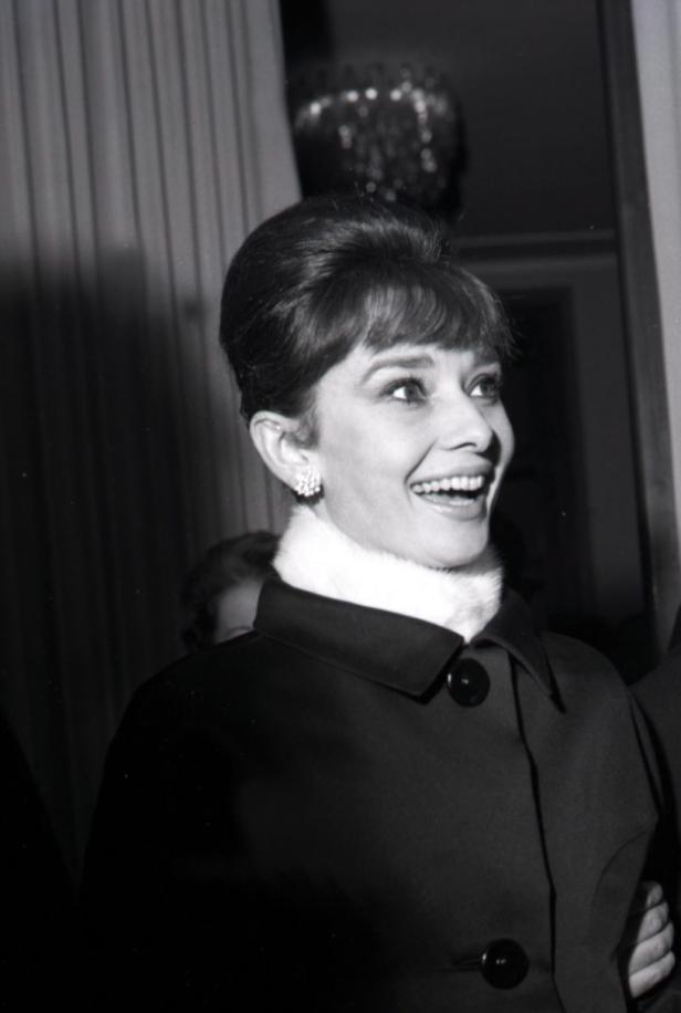 Audrey Hepburn lächelt in einem schwarzen Mantel mit weißem Kragen.