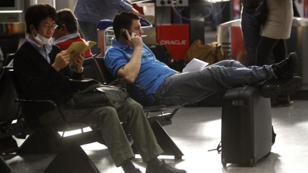 Passagiere sollen sich im Vorfeld bei ihrem Reiseveranstalter informieren. Zwei Männer warten in einem Flughafen, einer liest, der andere telefoniert mit Füßen auf einem Koffer.