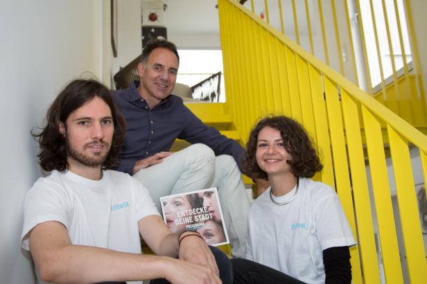 Drei Personen sitzen auf einer Treppe mit gelbem Geländer und halten eine Broschüre mit dem Titel „Entdecke deine Stadt“.