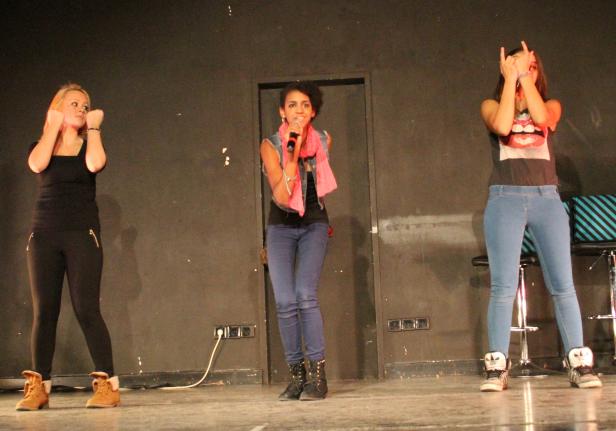 Drei junge Frauen stehen auf einer Bühne, eine singt ins Mikrofon.