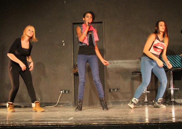 Eine Sängerin mit Mikrofon steht mit zwei Tänzerinnen auf einer Bühne.