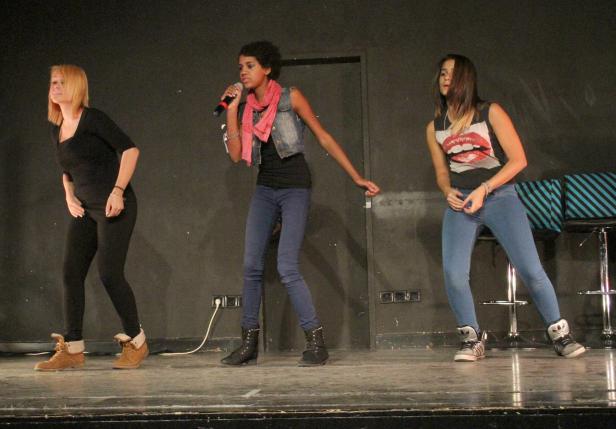 Drei junge Frauen stehen auf einer Bühne, die mittlere singt ins Mikrofon.