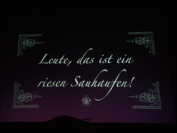 Der Text „Leute, das ist ein riesen Sauhaufen!“ wird auf einer Leinwand angezeigt.