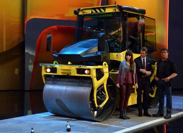 Eine gelbe Straßenwalze mit drei Personen davor auf einer Bühne.