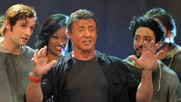 Sylvester Stallone gestikuliert auf einer Bühne, umgeben von anderen Darstellern.