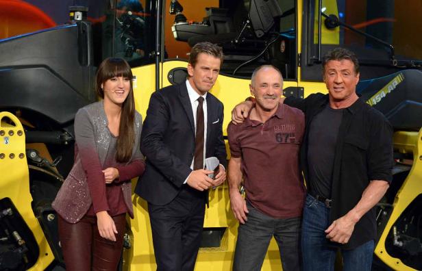 Sylvester Stallone und andere Personen posieren vor einer gelben Baumaschine.