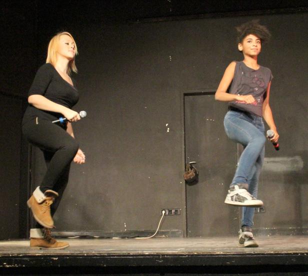 Zwei junge Frauen stehen mit Mikrofonen auf einer Bühne und tanzen.