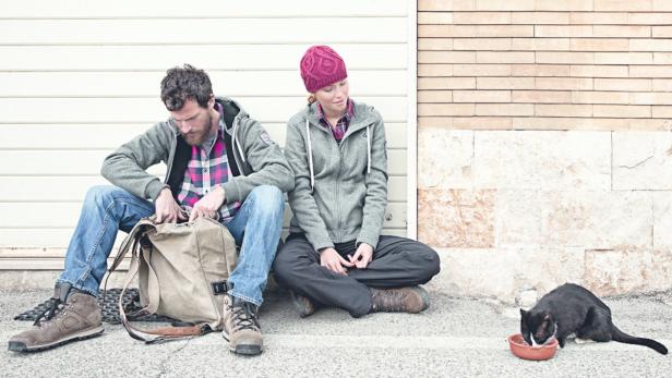 Ein Mann kramt in einem Rucksack, während eine Frau daneben sitzt und eine Katze frisst.
