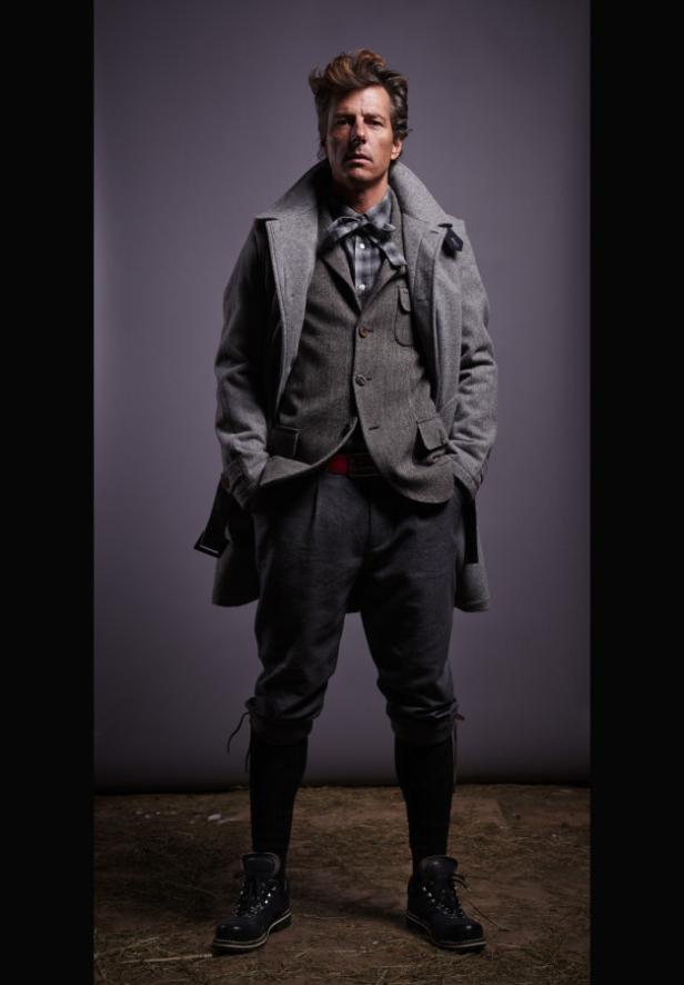 Ein Mann in einem grauen Tweed-Anzug, einem Mantel und Stiefeln posiert vor einem grauen Hintergrund.