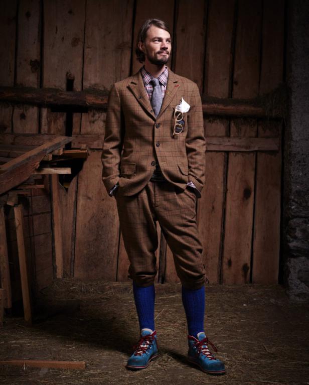 Ein Mann in einem Tweedanzug mit Kniebundhosen, blauen Socken und Wanderschuhen steht vor einer Holzwand.