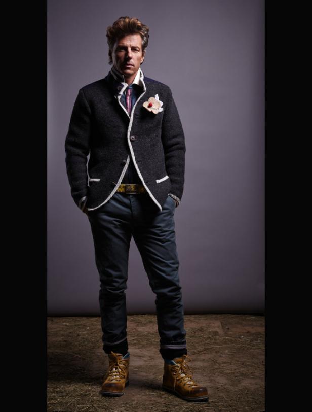 Ein Mann in Trachtenjacke, Jeans und Wanderstiefeln posiert vor einem grauen Hintergrund.